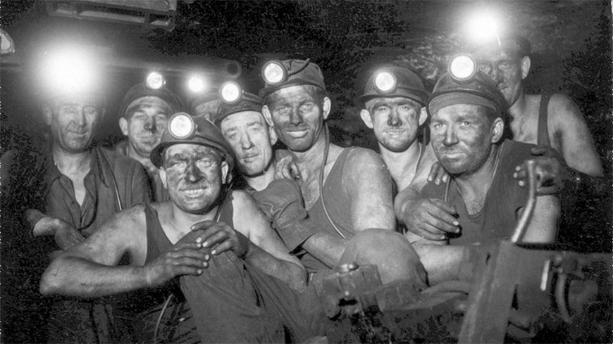 Photographie en noir et blanc d’un groupe de mineurs au fond de la mine, le visage marqué par le charbon et portant leur lampe sur leur casque.